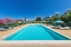Una piscina rectangular con agua azul cristalina, rodeada de sillones de descanso y sombrillas, y bordeada por una terraza de madera.