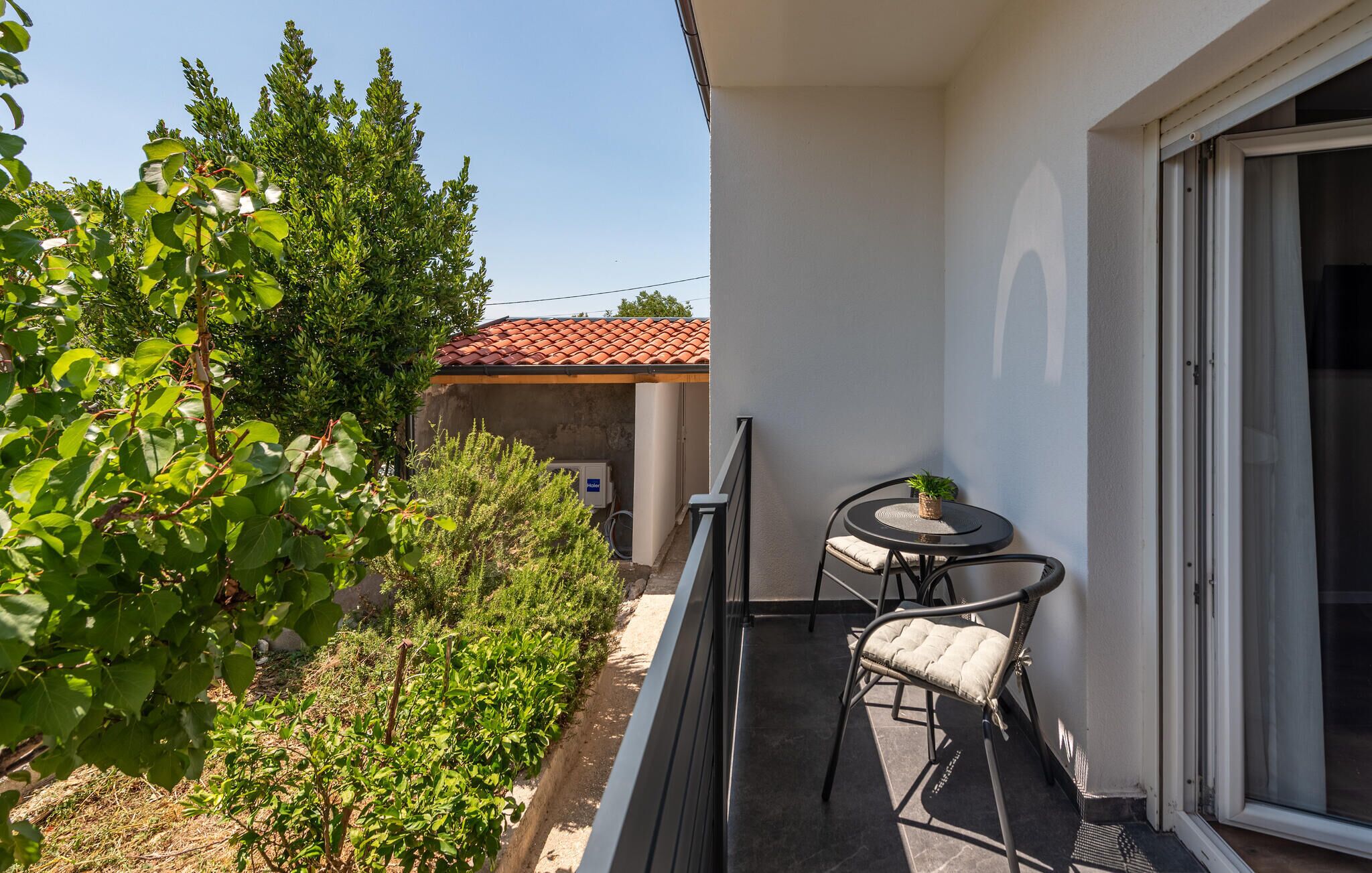 A balcony with a table and chairs, a potted plant, and a view of a building with a red-tiled roof.