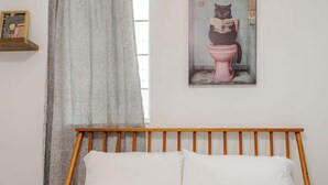 A bedroom with a wooden headboard, a bed with white bedding, a window with sheer curtains, and a wall-mounted picture of a cat reading a newspaper on a toilet. - Mins to Fishtown & Center City | GlobeNest (Philadelphia)