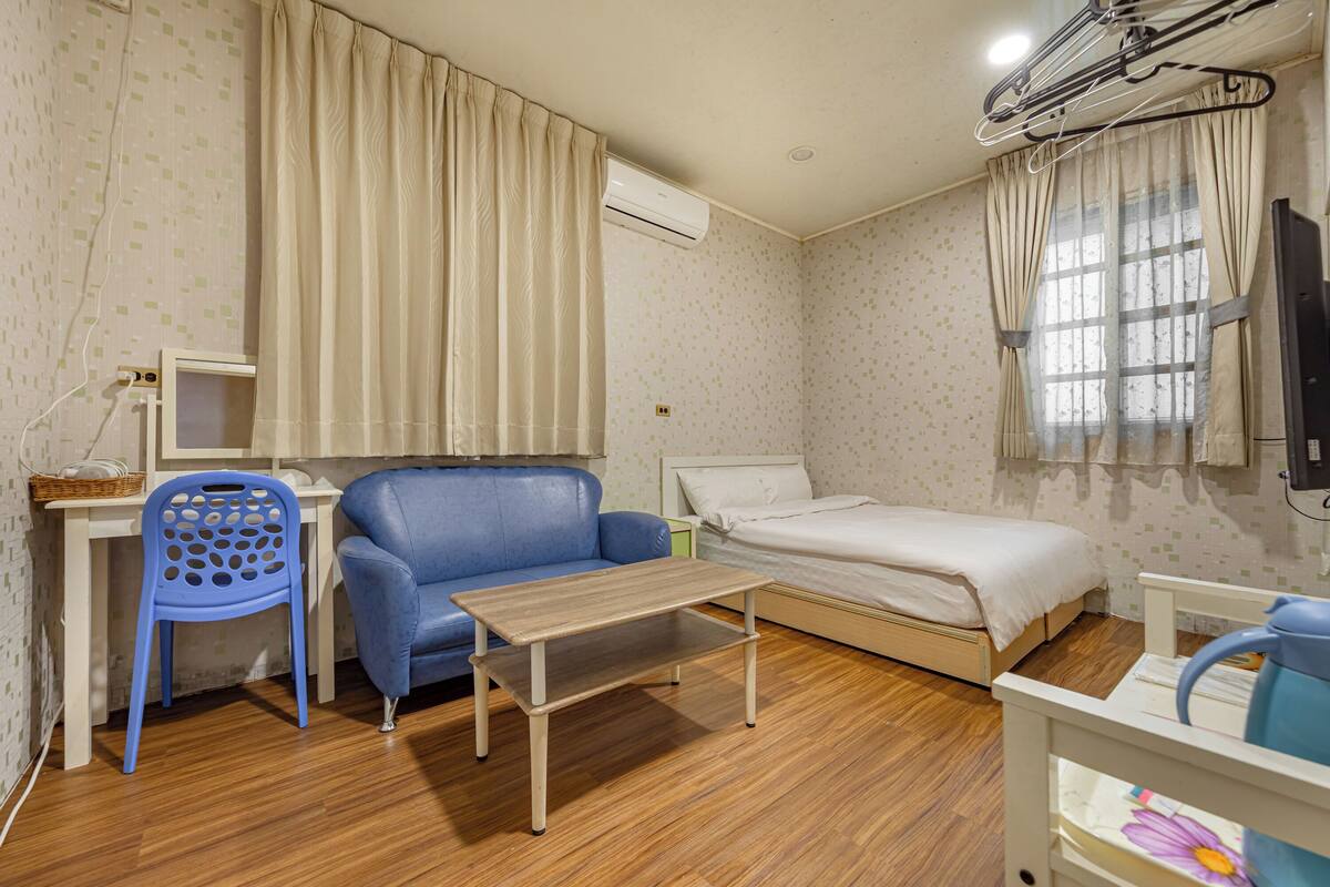 A small, well-lit room with a single bed, a blue chair, a wooden table, and a TV.