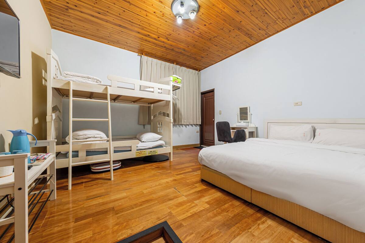 A room with a bunk bed, a single bed, wooden flooring, a desk with a chair, and a window with curtains.