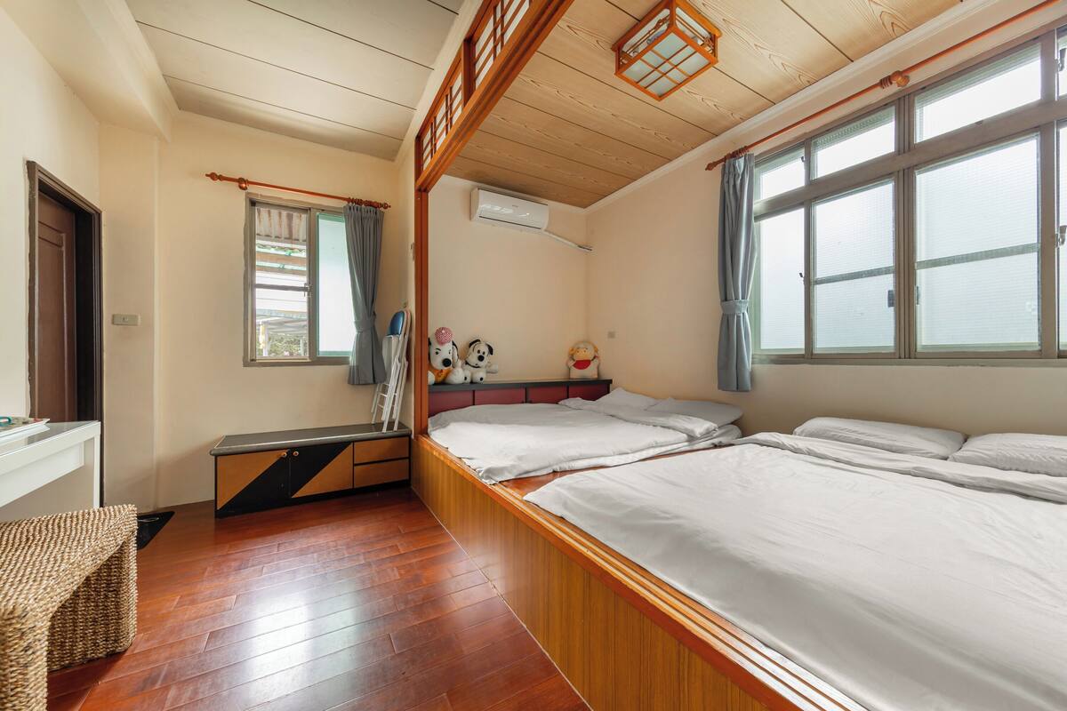 A room with a wooden floor, a bed with white bedding, a bench, and a window with a curtain.