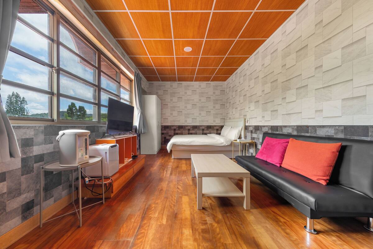 A modern room with a wooden ceiling, a bed, a black sofa with red cushions, a television, and a kitchen area with appliances.