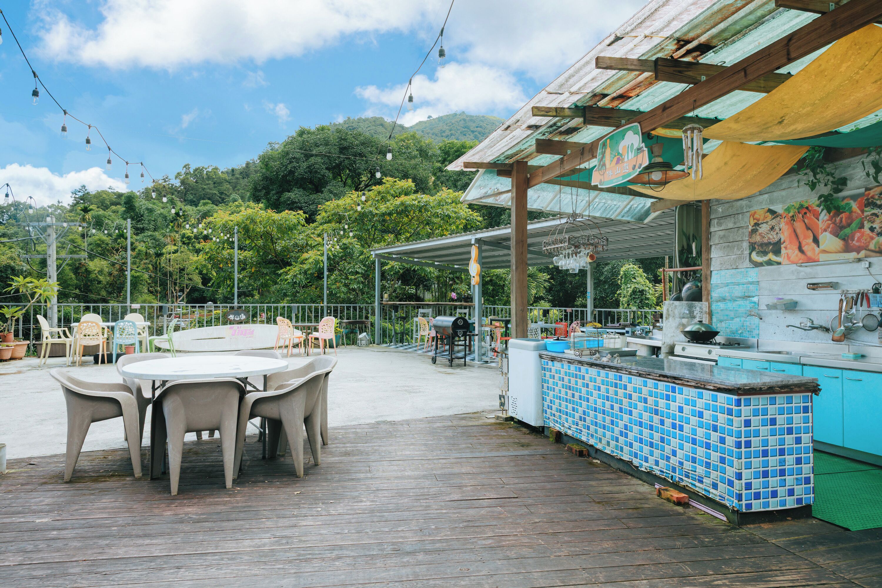 An outdoor patio area with a wooden deck, a round table with chairs, a bar area with blue tiles, and a view of a mountainous landscape.