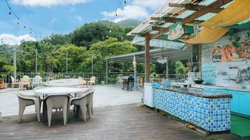 An outdoor patio area with a wooden deck, a round table with chairs, a bar area with blue tiles, and a view of a mountainous landscape.