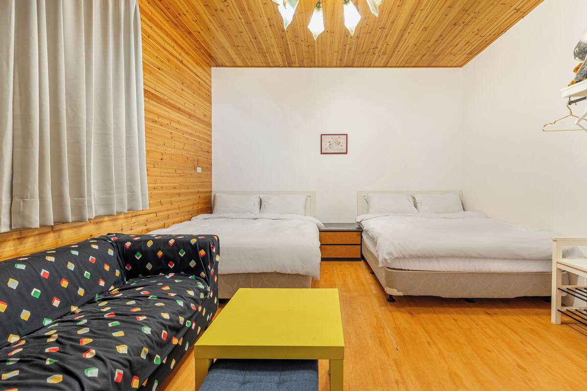 A room with two beds, a sofa, a small table, wooden flooring, and a wooden ceiling.