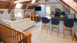 A cozy attic-style living room with a sofa, dining table, and chairs. A kitchen with appliances and cabinets is visible in the background. - Pendre Barn (Llanfyrnach)