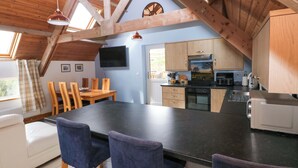 A modern kitchen with wooden beams, a dining area with a black table and blue chairs, and a TV mounted on the wall. - Pendre Barn (Llanfyrnach)