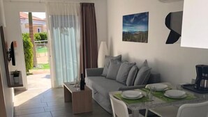 A modern living room with a grey sofa, white chairs, a dining table set for two, and a kitchen area with a stove. - Alsol Los Leones bungalows - Adults Only (San Bartolomé de Tirajana)