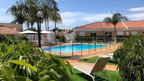 A swimming pool with a tiled deck, surrounded by palm trees and a building with a red-tiled roof. - Alsol Los Leones bungalows - Adults Only (San Bartolomé de Tirajana)