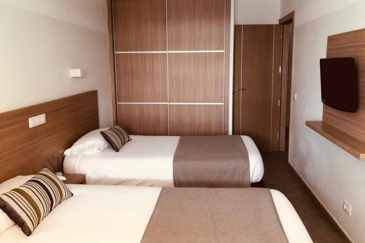 A hotel room with two beds, a wooden wardrobe, a television on a shelf, and a bedside table with a lamp.