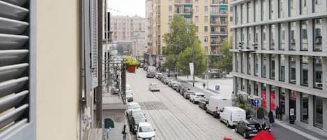 A street view from a balcony with a row of parked cars, a tram line, and buildings in the background.