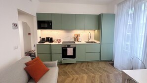 Cocina pequeña con gabinetes verdes, un microondas, una estufa y un lavamanos. El piso es de madera con un patrón de espiga y hay un sofá gris con un cojín naranja.