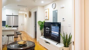 Ein modernes Wohnzimmer mit Flachbildfernseher, Glastisch und Essbereich, im Hintergrund ist eine Küche zu sehen.