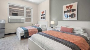 A hotel room with two beds, a kitchenette, a window with an air conditioner, and artwork on the walls.