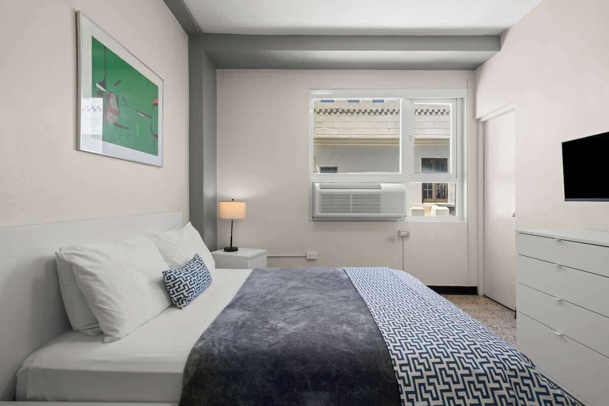 Une chambre à coucher comprenant un lit, une table de chevet avec une lampe, une fenêtre avec des stores et un téléviseur fixé au mur.