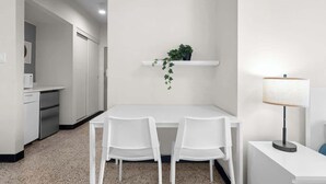 A modern kitchen with white cabinetry, a countertop, and a sink. There is a white dining table with two chairs and a small potted plant on a shelf above. A lamp is on a side table next to the dining area.