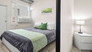A bedroom with a bed, a nightstand, a window, and a wall-mounted air conditioner. - With Balcony | King Bed |  Free Parking | AC (San Juan)