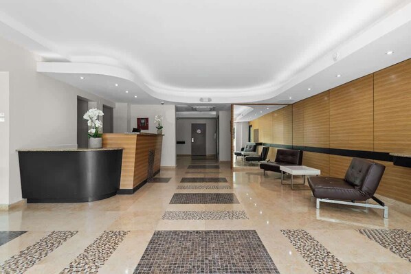 A modern hotel lobby with a reception desk, seating area, and a hallway leading to other rooms.