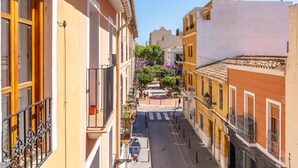 Una calle estrecha flanqueada por edificios, con balcones y un árbol en el centro con flores moradas.