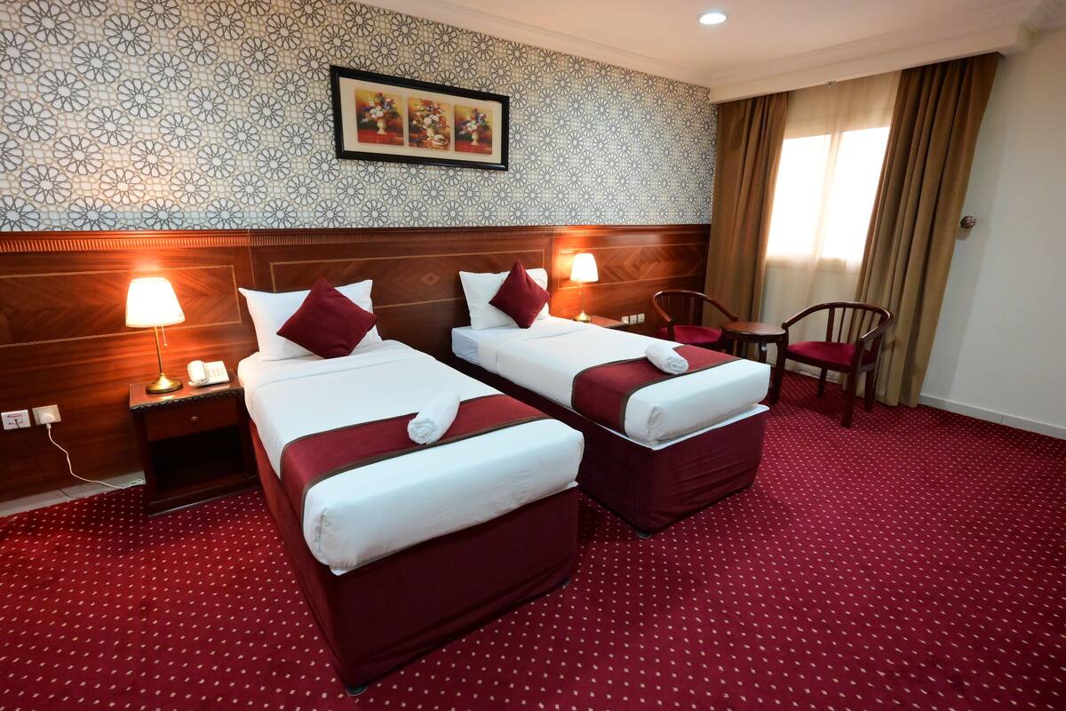Hotellihuone, jossa on kaksi sänkyä, puinen pääty, punainen matto, pieni pöytä ja tuoli.