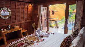 A cozy room with a wooden ceiling, a bed with white linens and rose petals, a small table with a kettle, and a view of a wooden house and garden through an open door. - Sayong Wooden House (Ubud)