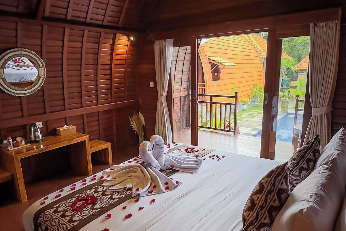 A cozy room with a wooden ceiling, a bed with white linens and rose petals, a small table with a kettle, and a view of a wooden house and garden through an open door.