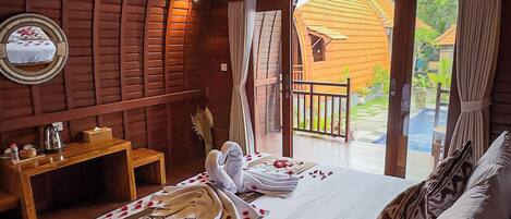 A cozy room with a wooden ceiling, a bed with white linens and rose petals, a small table with a kettle, and a view of a wooden house and garden through an open door.