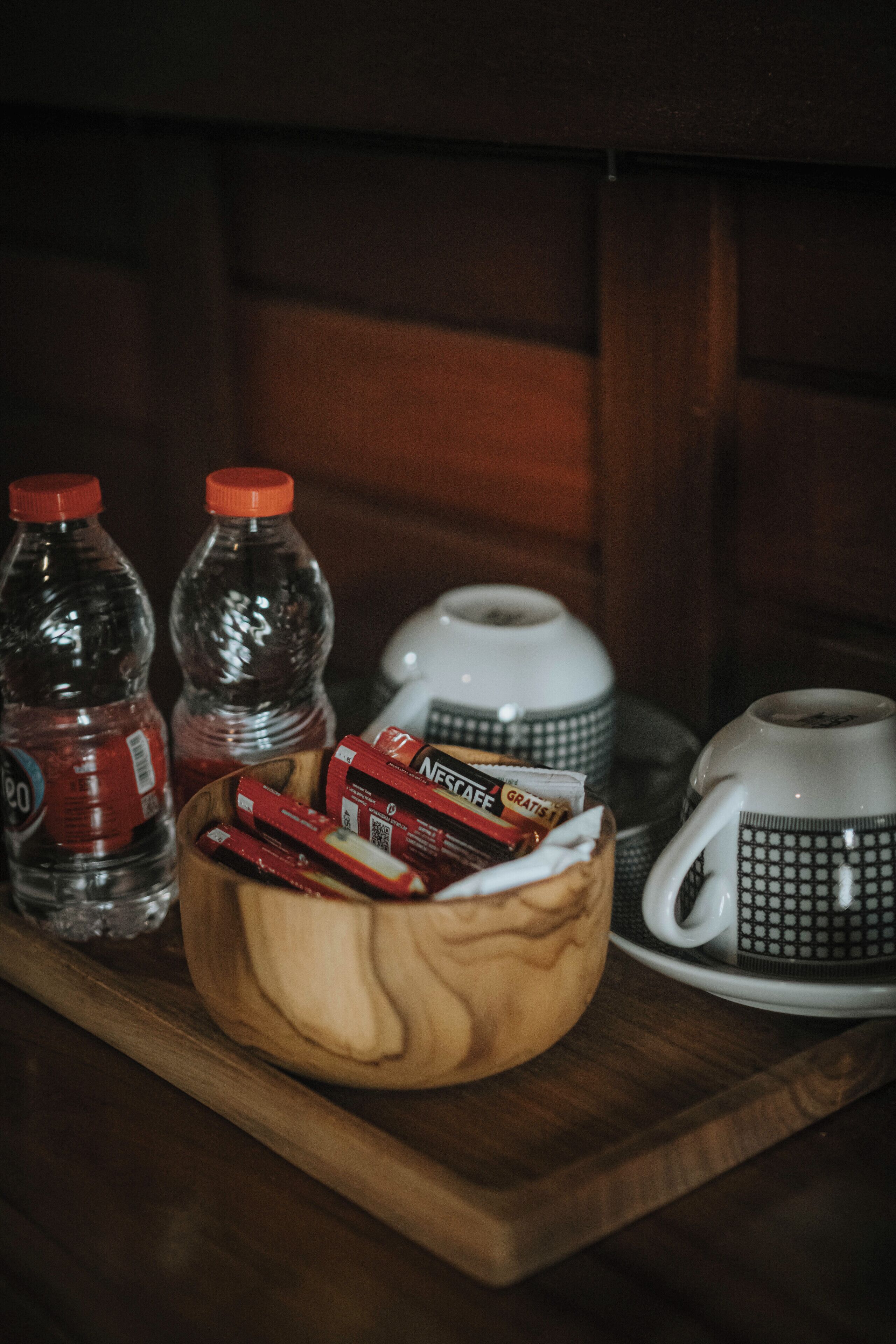 A wooden tray with two bottles, a wooden bowl with snacks, and two coffee cups.
