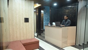 A reception area with a wooden reception desk, a red bench, and a staircase in the background. - Hotel Kumar Continental (Jhusi)