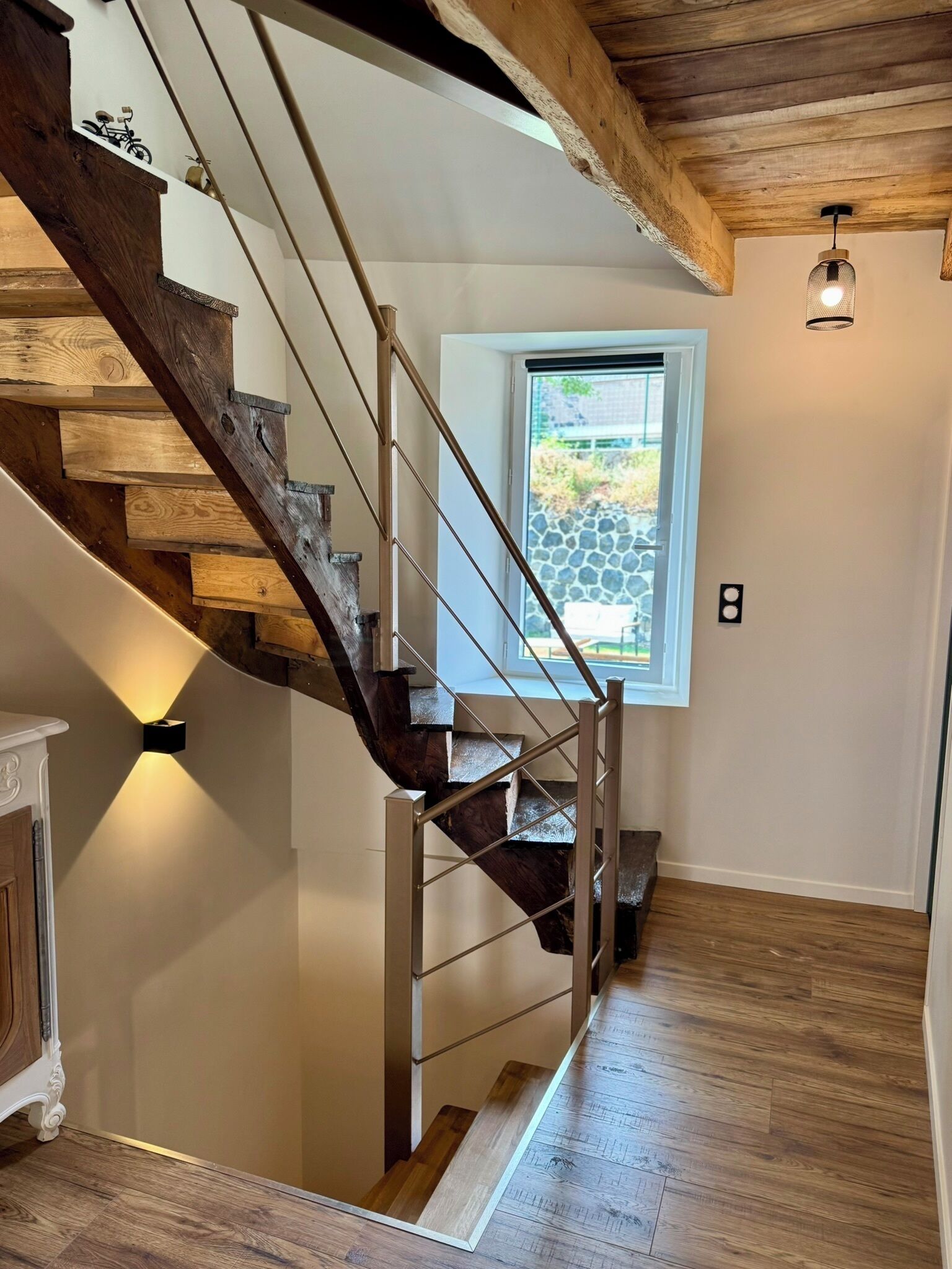 A wooden staircase with a metal railing, a small window with a stone wall view, and a wall-mounted light fixture.