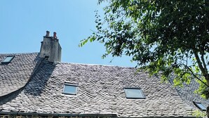 A stone house with a sloped roof, multiple windows with shutters, a small garden area, and a staircase leading to the entrance. - House in the shadow of the bell tower (Laguiole)
