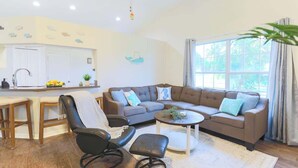 Una sala de estar moderna con un sofá marrón, un sillón de cuero negro, una mesa de centro y una zona de cocina con un armario blanco y un lavamanos.