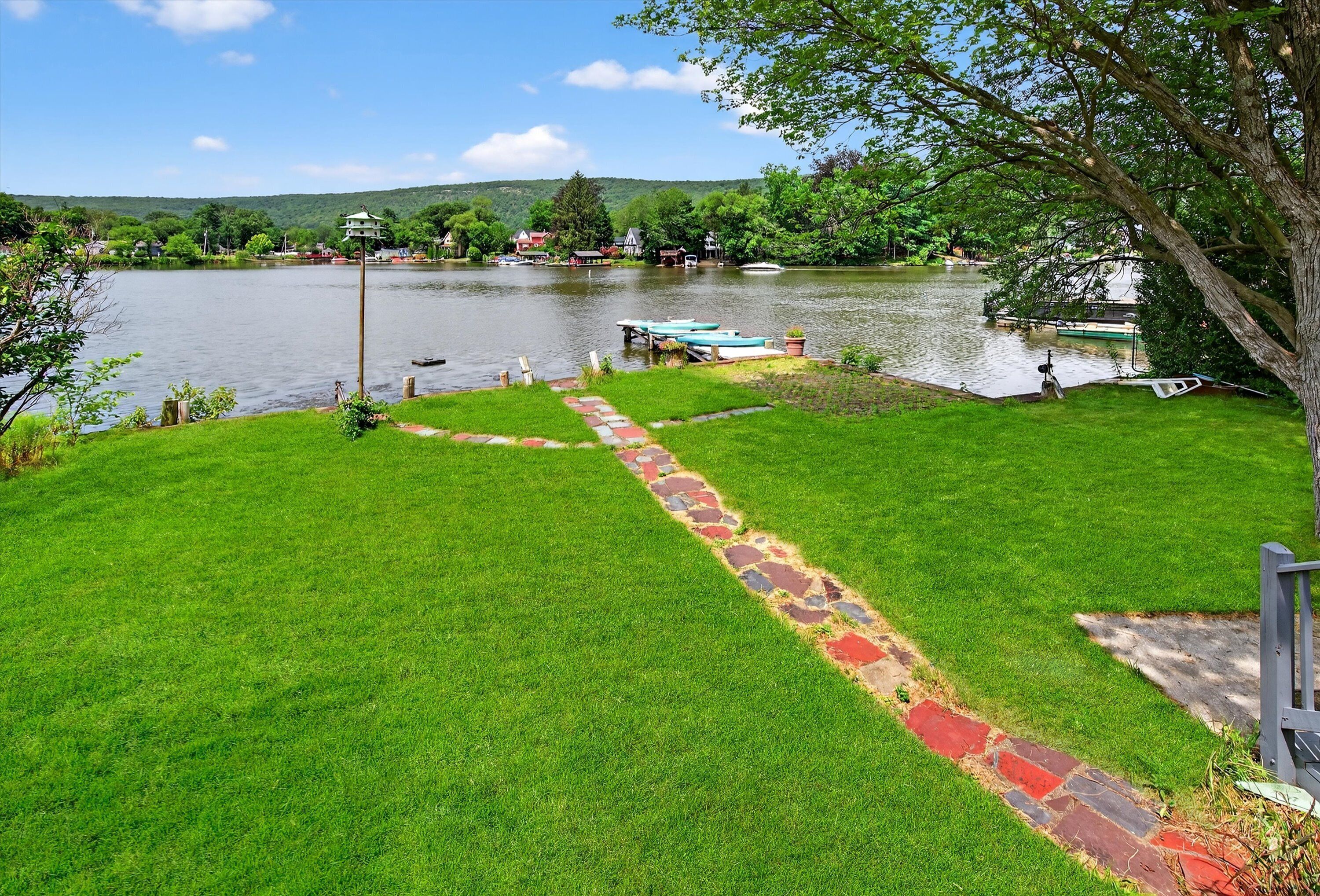 A grassy area with a stone pathway leading to a dock by a lake, surrounded by trees and houses in the background.