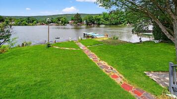 A grassy area with a stone pathway leading to a dock by a lake, surrounded by trees and houses in the background.