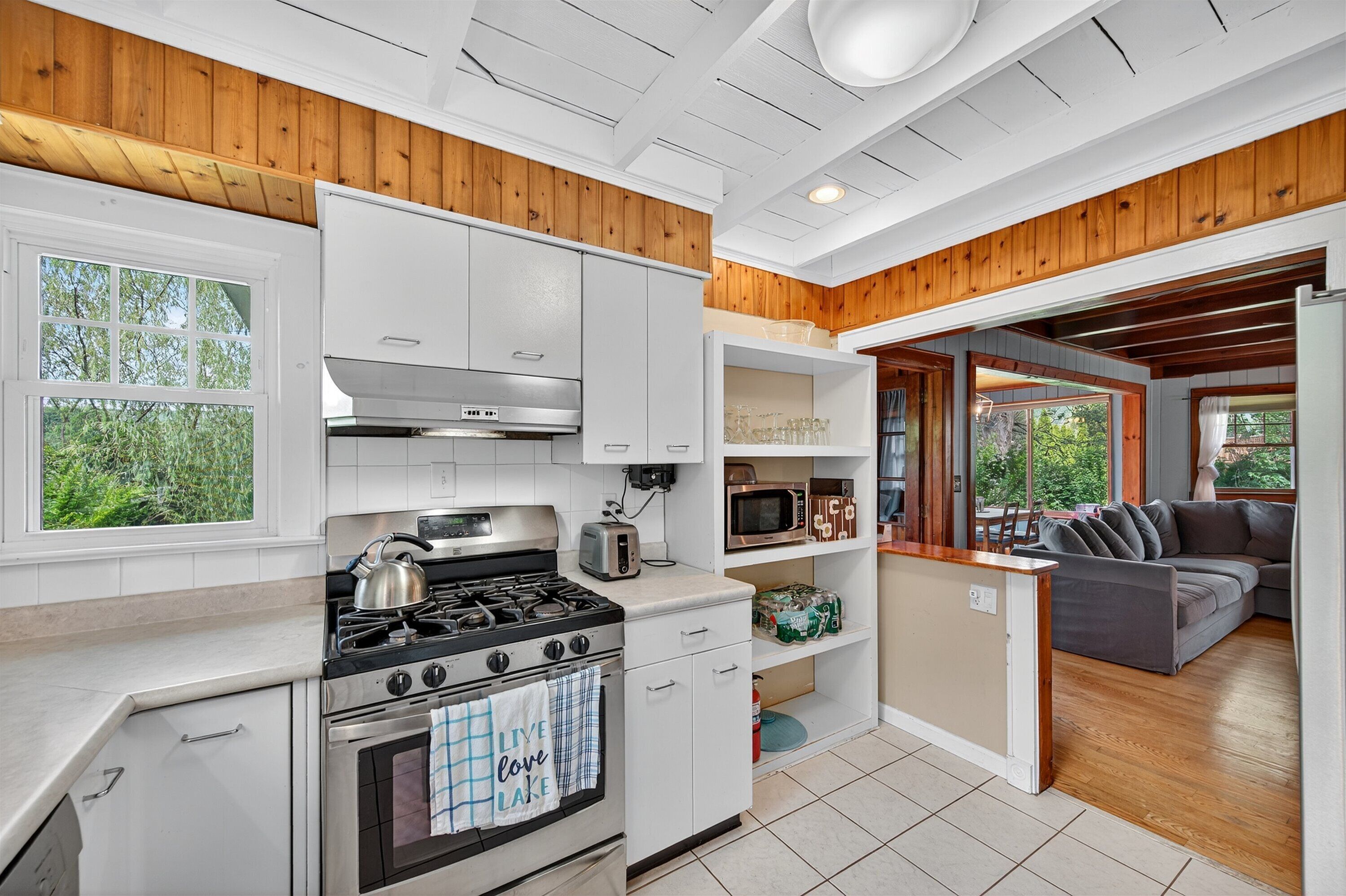 A modern kitchen with white cabinets, a stove, and a microwave. There is a wooden ceiling and a gray couch in the adjacent living area.