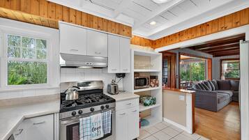A modern kitchen with white cabinets, a stove, and a microwave. There is a wooden ceiling and a gray couch in the adjacent living area.