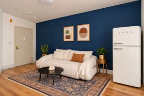 Un salon comprenant un canapé, une table basse, un tapis à motifs, un réfrigérateur blanc et deux tableaux encadrés accrochés au mur.