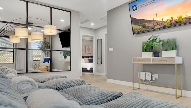 A modern hotel room with a large flat-screen TV, a sofa with rolled-up blankets, a console table with plants, and a view of a desert landscape. - 4BR w pool, fire pit, game room (Queen Creek)