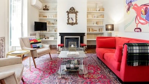 Una sala de estar con un sofá rojo, un sillón color beige, una mesa de centro y una chimenea. Hay estanterías y un televisor en la habitación.