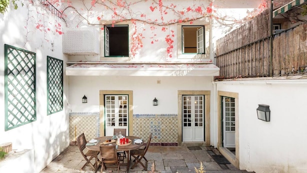 Un patio con una mesa de comedor dispuesta para cuatro personas, rodeada de sillas. Hay dos puertas y dos ventanas con contraventanas, y una valla de madera a la derecha.