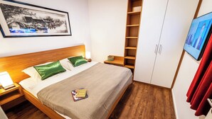 A hotel room with a wooden bed, green pillows, a framed picture on the wall, a wardrobe, and a TV on the wall.