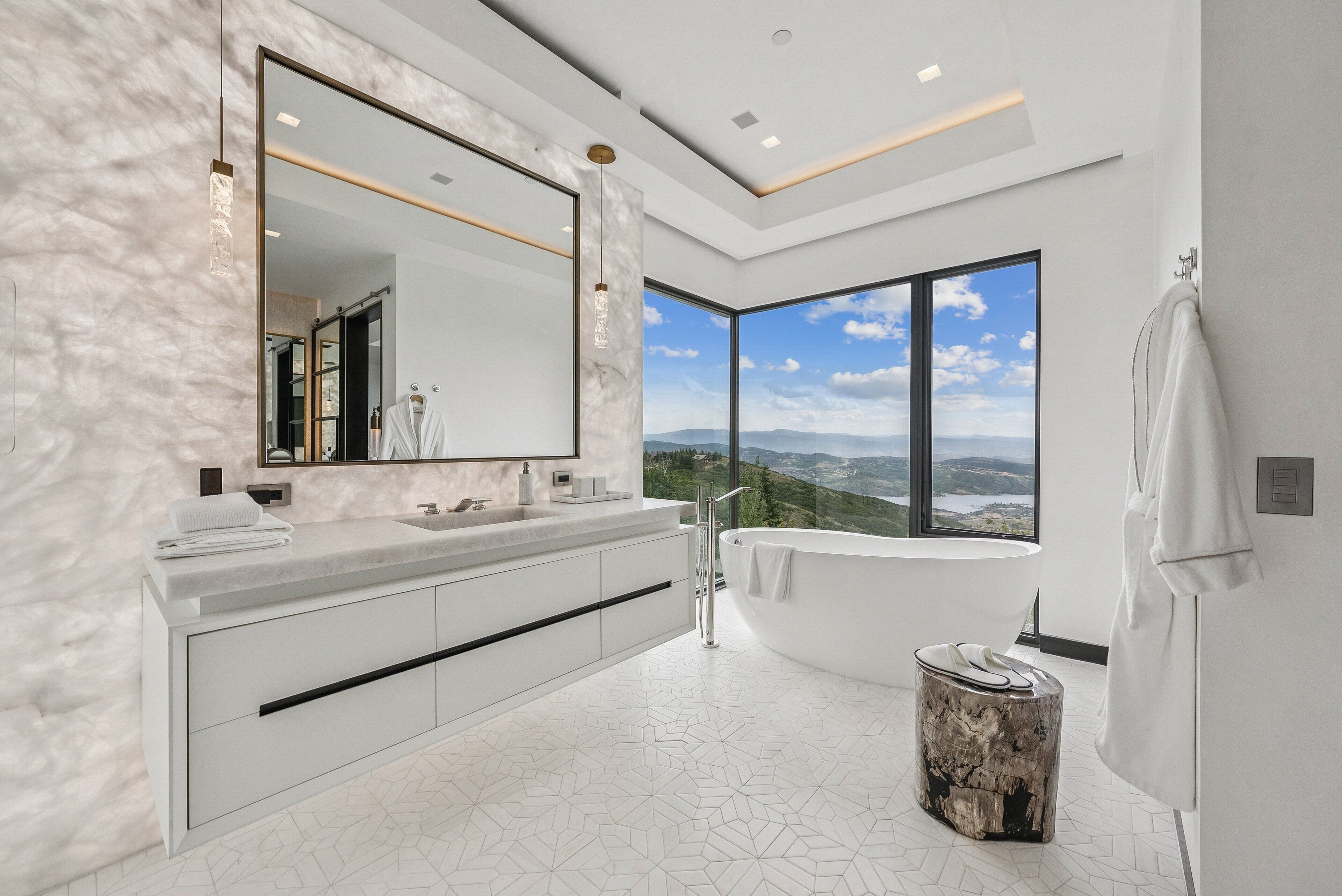 A modern bathroom with a large mirror, a freestanding bathtub, and a view of the mountains through large windows.