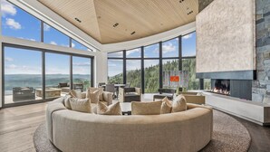 Una sala de estar moderna con un sofá circular grande, una chimenea y ventanas amplias con vistas a las montañas.