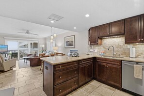 A kitchen with dark wooden cabinets, a large island with a sink, and a dishwasher. There is a dining area with a table and chairs visible through an open door.