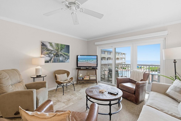 A living room with a television, comfortable seating, a ceiling fan, and a view of the ocean.