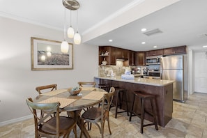 A modern kitchen with a central island, dark wooden cabinets, stainless steel appliances, and a dining area with a glass table and wooden chairs.