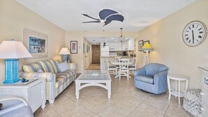 A living room with a sofa, armchair, and coffee table. There is a kitchen in the background with a dining table and chairs. - Beachfront Comfort w/ Pool, Balcony & Steps to Flagler Avenue (New Smyrna Beach)