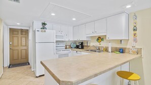 A kitchen with white cabinets, a central island with stools, a microwave, and a refrigerator. - Beachfront Comfort w/ Pool, Balcony & Steps to Flagler Avenue (New Smyrna Beach)