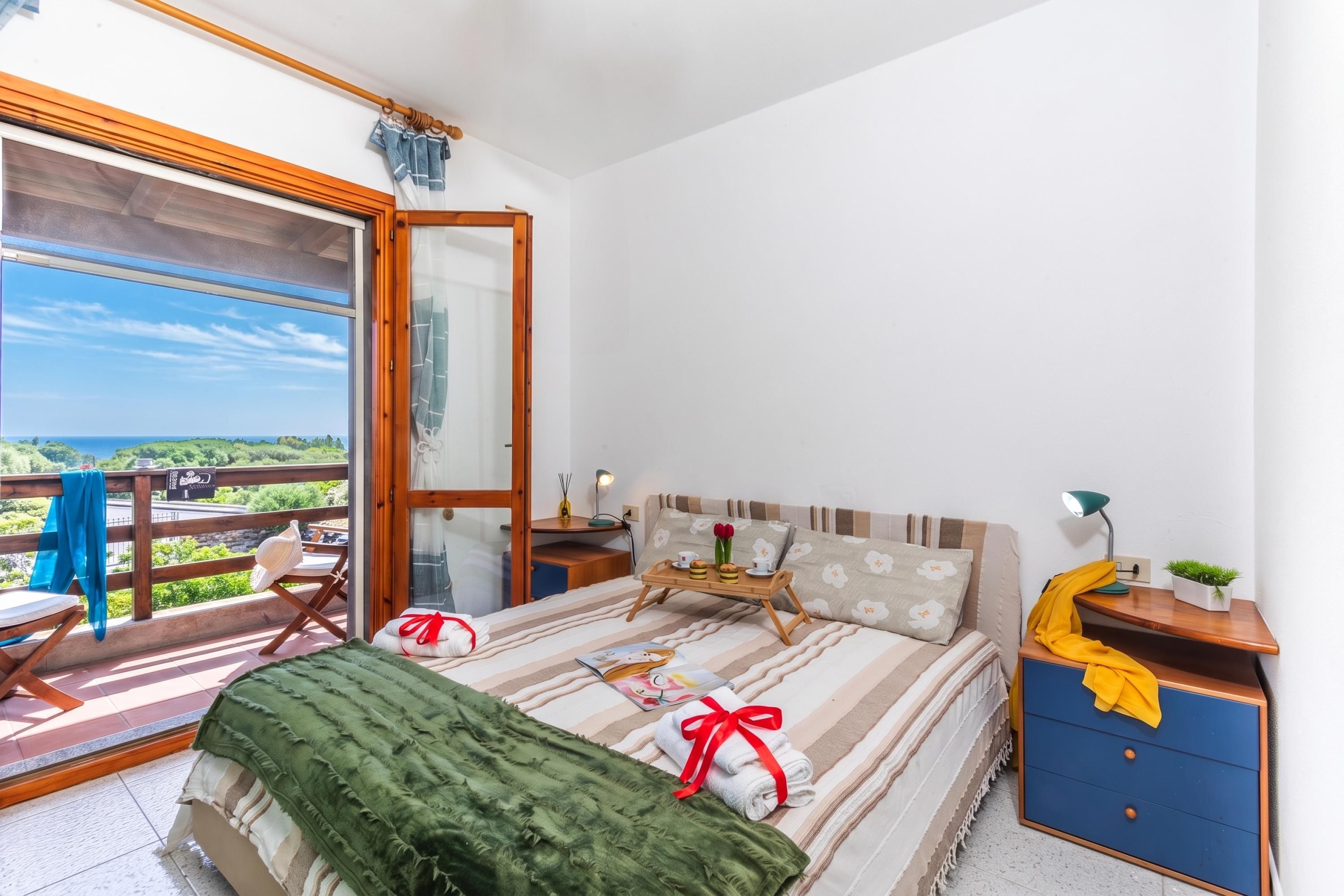 A bedroom with a bed, a bedside table, a nightstand, a window with a view, and a balcony with a chair.